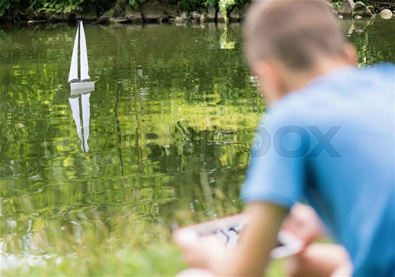 Teen boy playing with a remote controlled boat. Handmade model sailboat on lake - children is playing with tablet. Selective focus limited to boat, stock photo