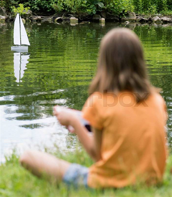 Girl playing with a remote controlled boat. Handmade model sailboat on lake - child is playing with tablet. Out of focus girl. Selective focus limited to boat, stock photo