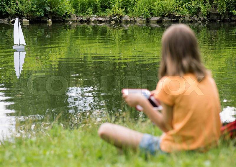 Girl playing with a remote controlled boat. Handmade model sailboat on lake - child with tablet. Out of focus girl. Selective focus limited to boat, stock photo