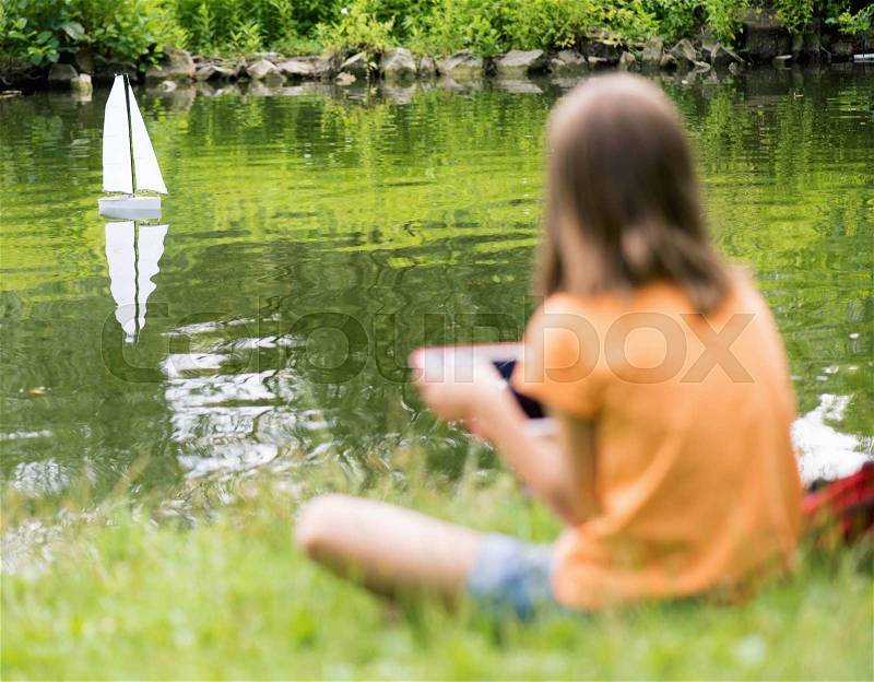 Girl playing with a remote controlled boat. Handmade model sailboat on lake - child is playing with tablet. Out of focus girl. Selective focus limited to boat, stock photo