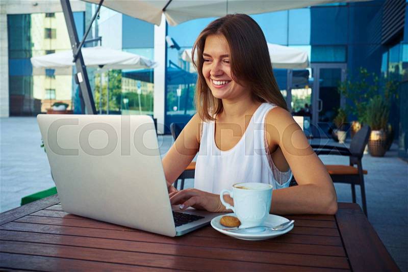Young businesswoman is sitting in outdoor café with a cup of coffee and working on her laptop against modern mirrored glass building background, stock photo