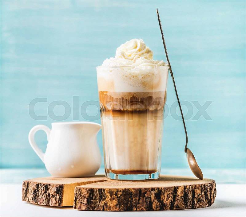Latte macchiato with whipped cream, serving silver spoon and pitcher on wooden round board over blue painted wall background, selective focus, horizontal composition, stock photo