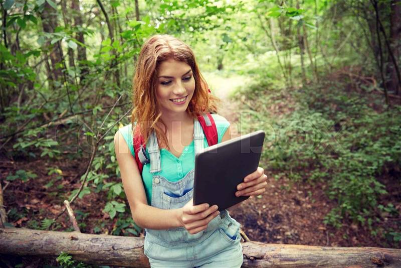 Adventure, travel, tourism, hike and people concept - happy young woman with backpack and tablet pc computer sitting on fallen tree trunk in woods, stock photo