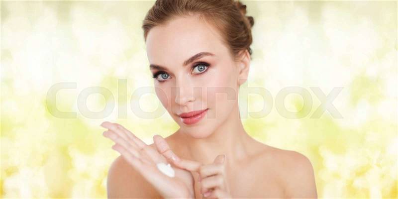 Beauty, people, skincare and cosmetics concept - happy young woman with moisturizing cream on hand over yellow holidays lights background, stock photo