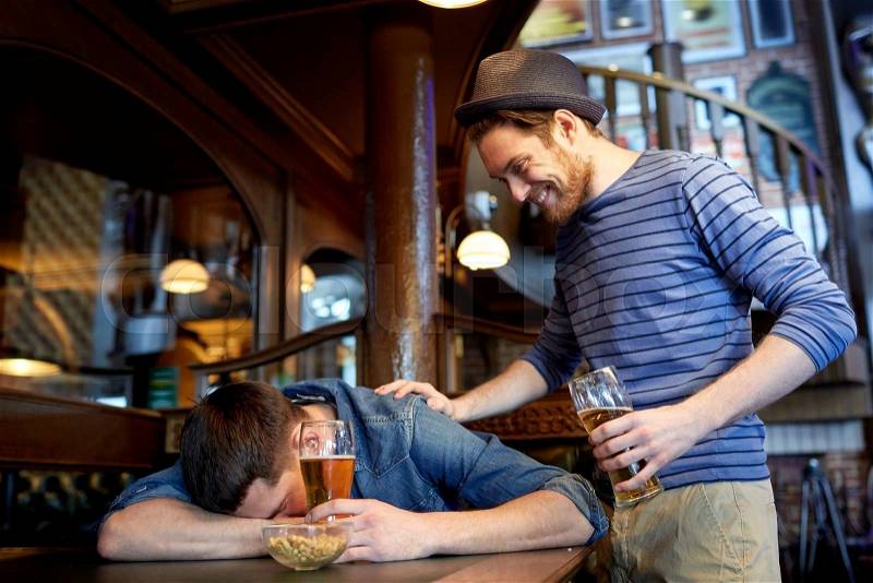 People, leisure, friendship and party concept - man with beer waking his drunk friend sleeping on table at bar or pub, stock photo