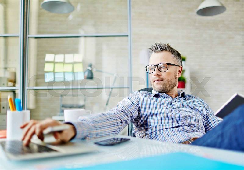 Modern businessman with touchpad sitting at workplace and looking at laptop display, stock photo