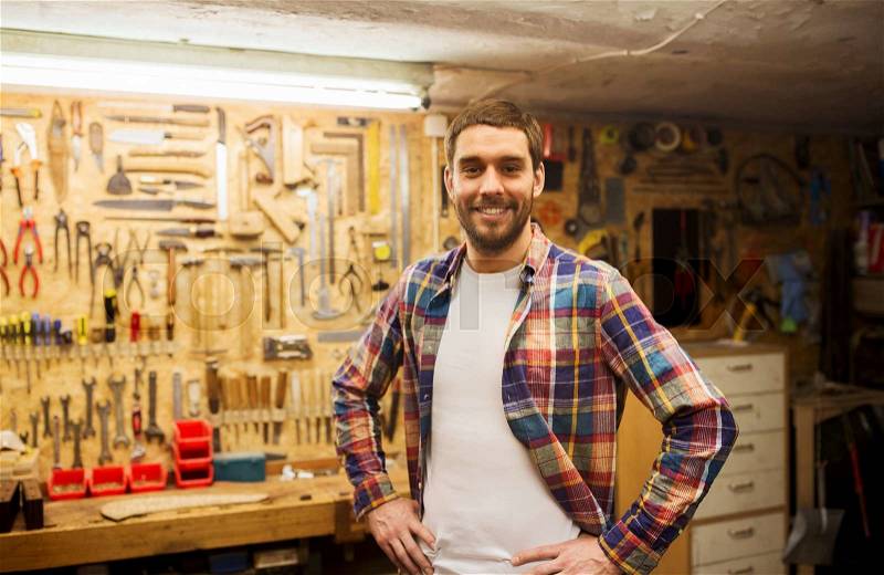 Profession, people, carpentry and people concept - happy man or carpenter in checkered shirt standing at workshop wall with work tools, stock photo
