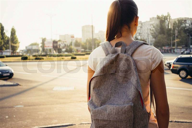 Slender young brunette in a shirt and a backpack in a city in the light of a sunset, view from the back, stock photo