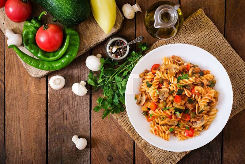 Vegetarian Vegetable pasta Fusilli with zucchini, mushrooms and capers in white bowl on wooden table. Top view, stock photo