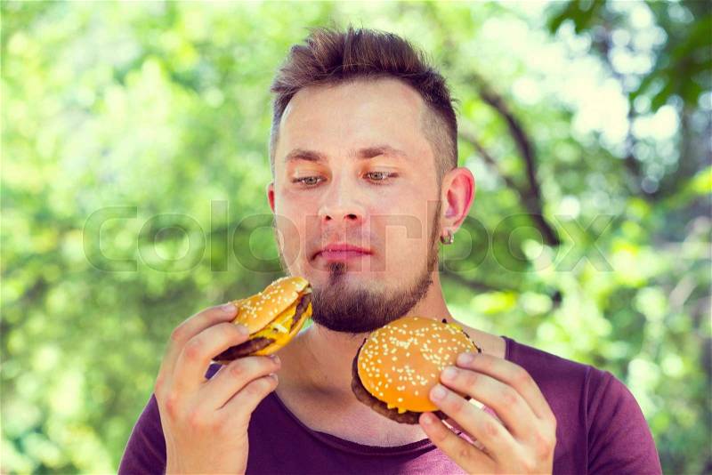 Emotional young guy eating a cheeseburger on the nature, stock photo