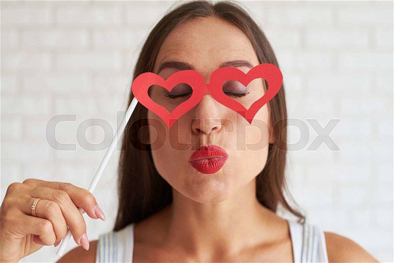 Smiling brunette holding red glasses-mask in the shape of heart and kissing something, white brick wall on background, stock photo