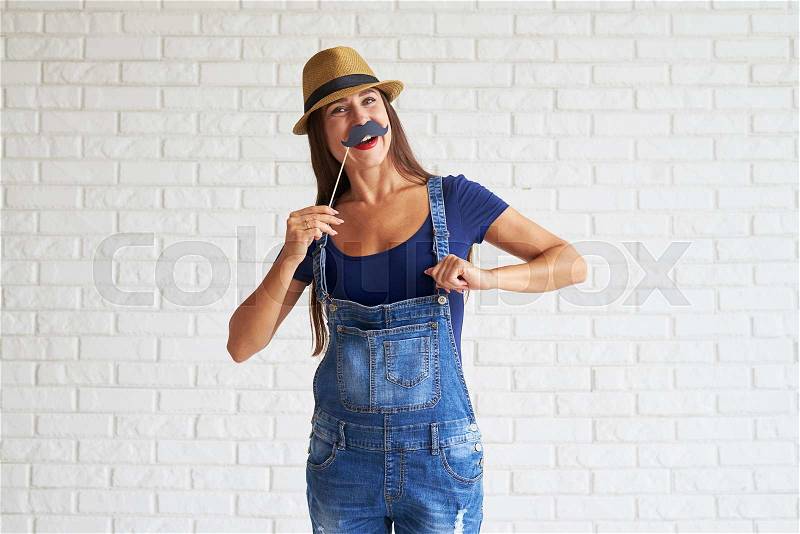 Beautiful happy woman wear denim overalls with straw hat and holding mustache mask, white brick wall on background, stock photo