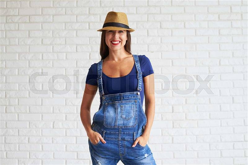 Beautiful happy women wear denim overalls and holds her hands in pockets, white brick wall on background, stock photo