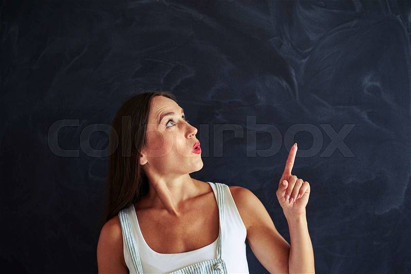 Pretty young woman is looking and pointing her finger up, black desk in background, stock photo