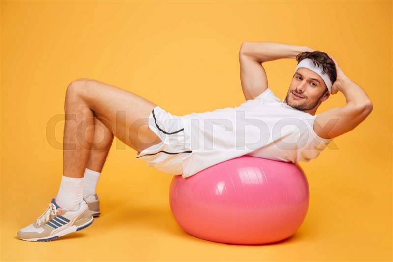 Young handsome sportsman doing abs on fitness ball isolated on the orange background, stock photo