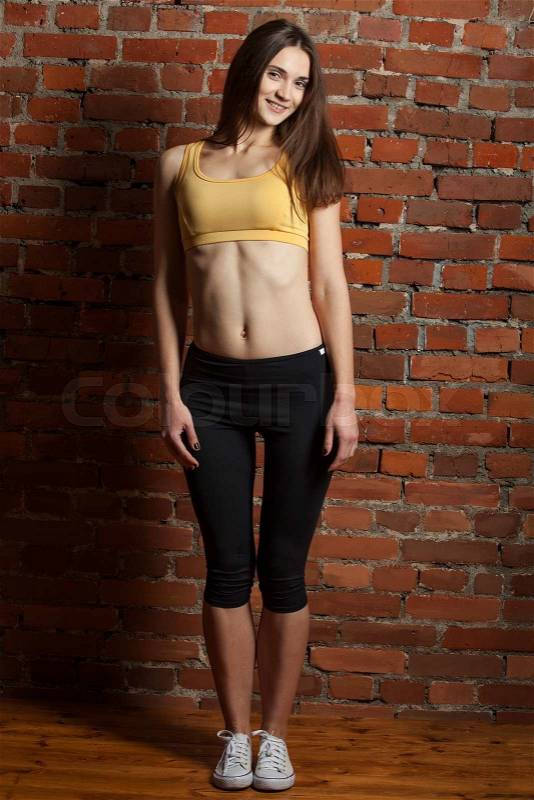 Young and attractive woman in top sports and leggings costs about red brick wall and smiles enigmatically, stock photo