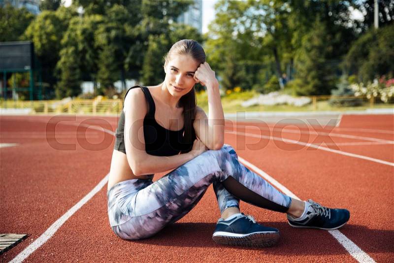 Fitness young woman sitting on running track after workout at the stadium, stock photo