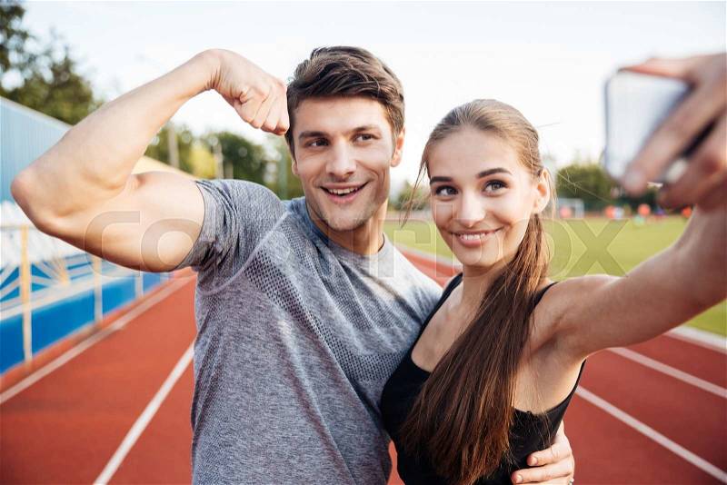 Young happy sports couple making selfie photo on stadium, man showing biceps on camera, stock photo