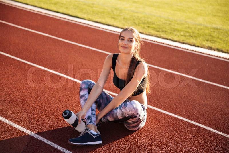 Fitness young woman sitting on running track and holding water bottle at the stadium, stock photo