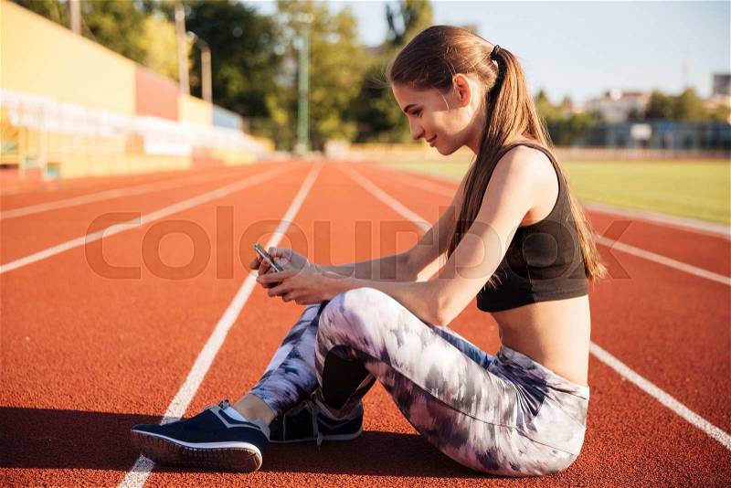 Happy female athlete using mobile phone on running track at the stadium, stock photo