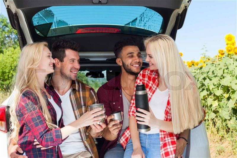 Friends drinking tea holding coffee cups thermos sitting in car trunk outdoor countryside, happy smile people group summer sunflower field, stock photo