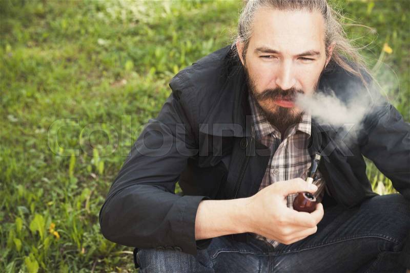 Asian man smoking a pipe on green grass in park, closeup photo with selective focus, stock photo
