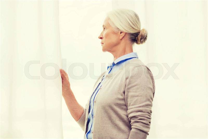 Age, loneliness and people concept - lonely senior woman looking through window at home, stock photo