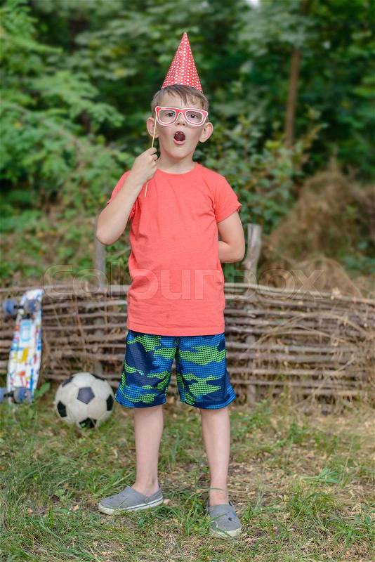 Happy young boy playing with photo booth props or accessories standing in the garden with his football holding a pair of glasses to his eyes and wearing a party hat, stock photo