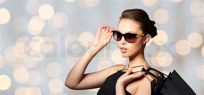 Sale, fashion, people and luxury concept - happy beautiful young woman in black sunglasses with shopping bags over holidays lights background, stock photo