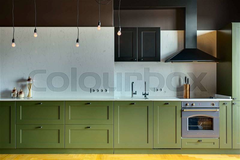 Kitchen in a modern style with a light tabletop with sink, cooker, oven, kitchen accessories. Under tabletop there are green drawers. Over tabletop there is kitchen hood, cupboard and glowing lamps, stock photo