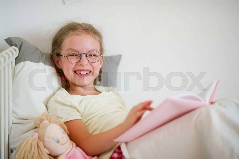 A fun girl in glasses and pigtails reads a book while lying in bed. Next to her sits a doll. The girl looks into the camera laughing, stock photo