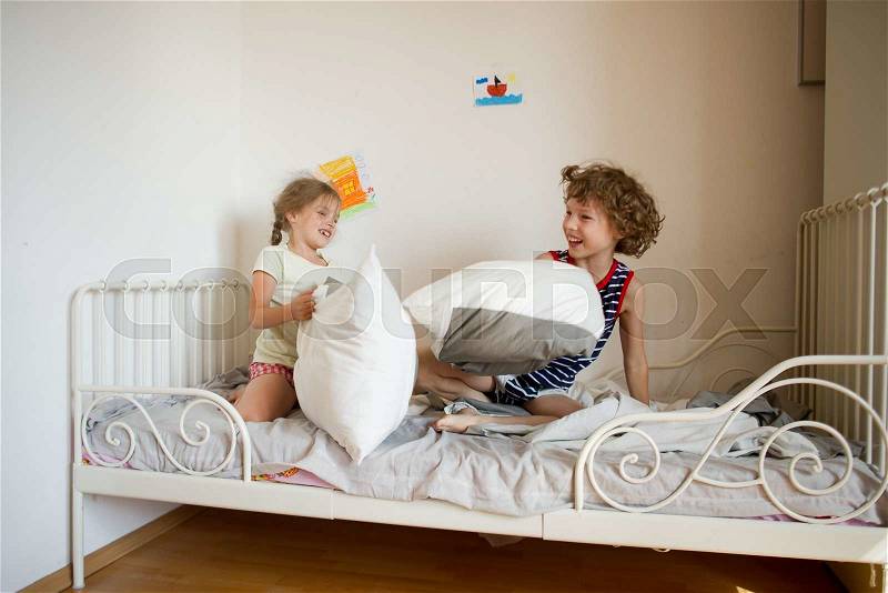Little brother and sister staged a pillow fight in the bedroom. They sit on a bed in pajamas. Children hair uncombed. Happy smiles on their faces, stock photo