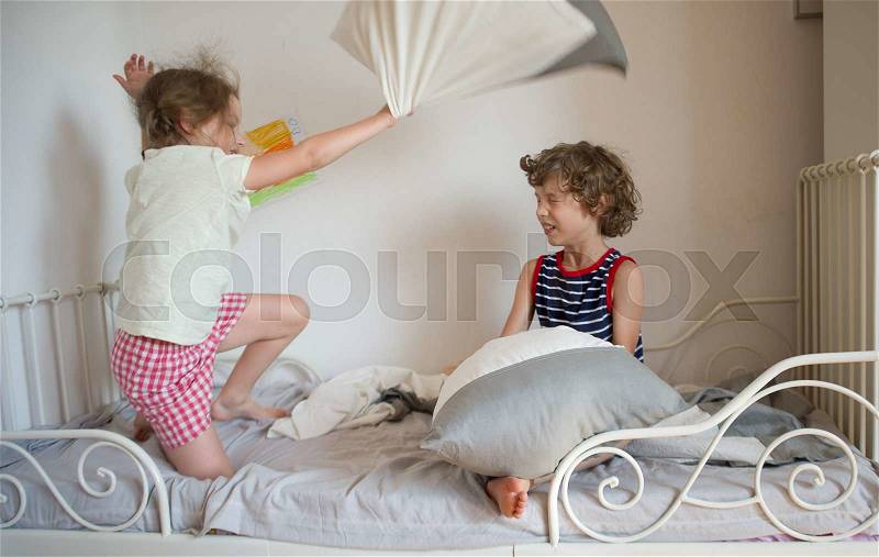The brother and the sister have arranged fight by pillows on a bed in a bedroom. The naughty little girl beats the brother a pillow. The boy has closed eyes. Children love this game, stock photo