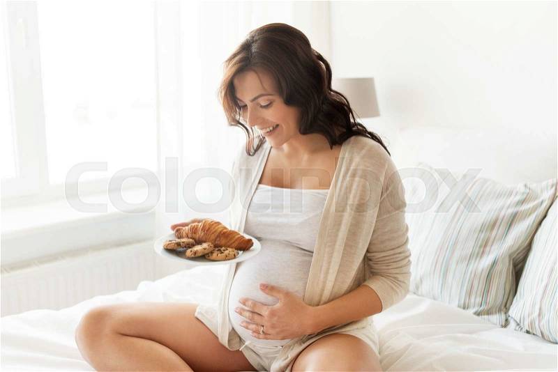 Pregnancy, unhealthy eating, food and people concept - happy pregnant woman with croissant buns at home, stock photo