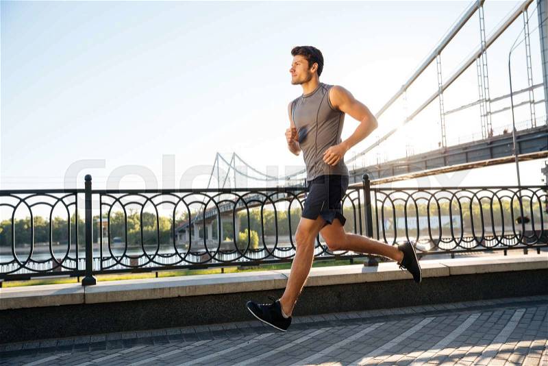 Side view of a handsome sports man running fast along big modern bridge at sunset light, stock photo