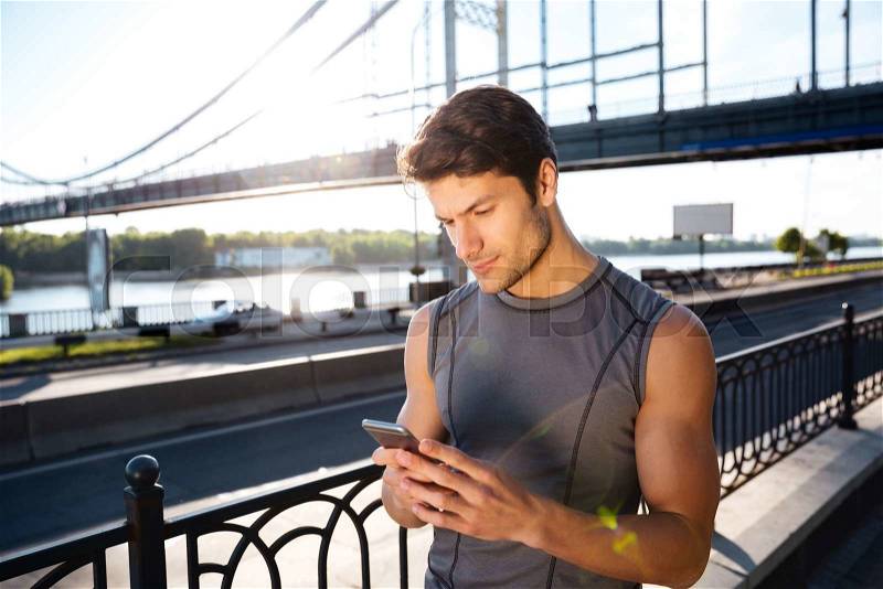 Male runner typing something on his mobile smart phone and standing at sunset on the bridge, stock photo