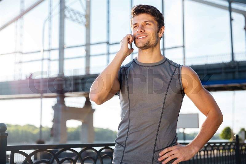 Handsome young man in sports uniform is talking on the mobile phone while standing at the bridge, stock photo