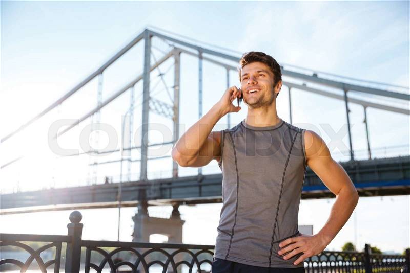 Handsome smiling young man in sports uniform is talking on the mobile phone while standing at the bridge, stock photo