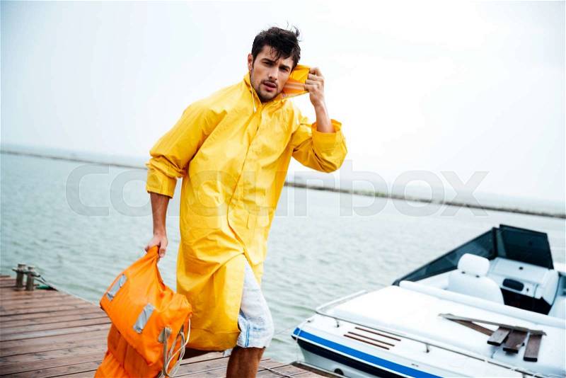Young handsome sailor man in yellow cloak walking at the sea pier holding life vest, stock photo