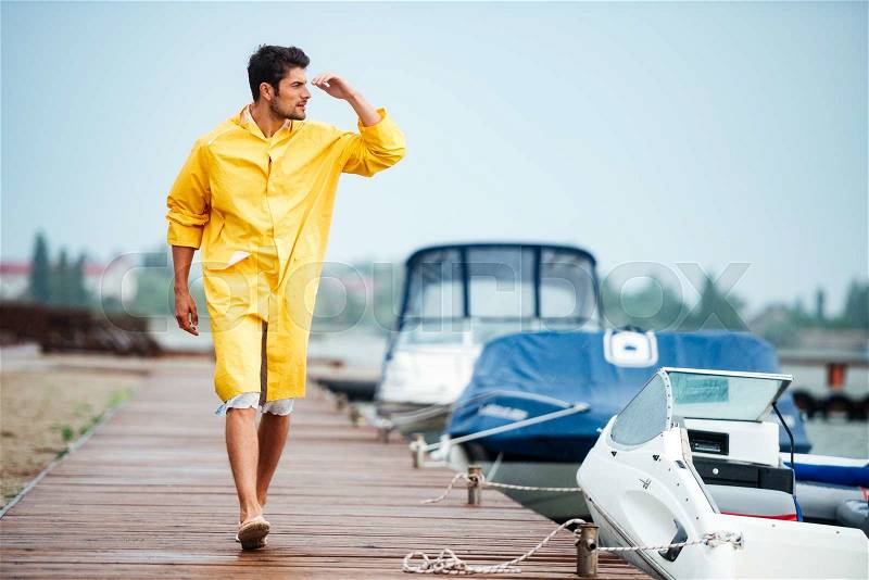 Young handsome sailor man in yellow cloak walking at the sea pier and looking at something, stock photo
