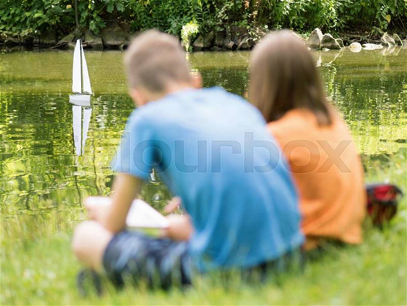 Girl and boy playing with a remote controlled boat. Handmade model sailboat on lake - children is playing with tablet. Selective focus limited to boat, stock photo