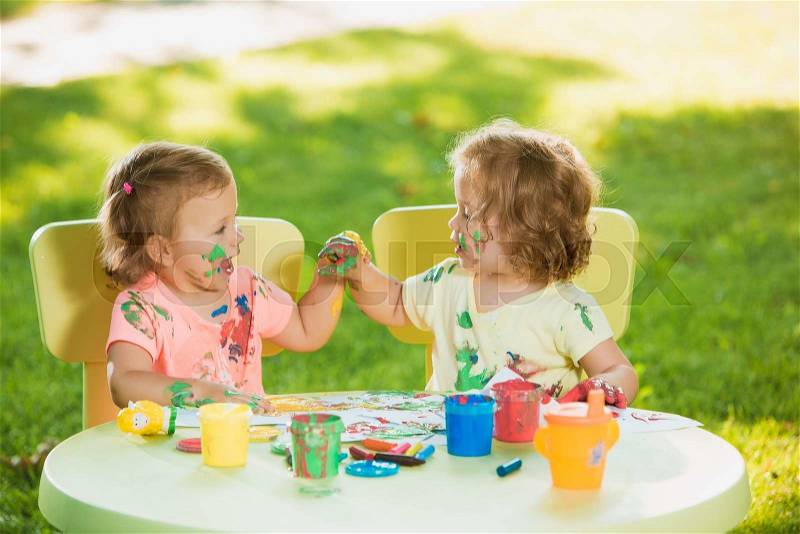 Two-year old girls painting with poster paintings and sitting at a table together against green lawn, stock photo