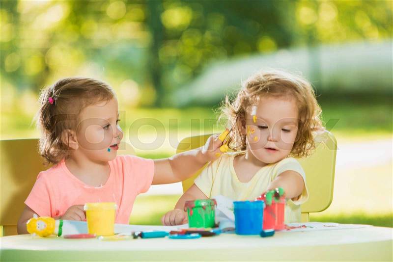 Two-year old girls painting with poster paintings and sitting at a table together against green lawn, stock photo