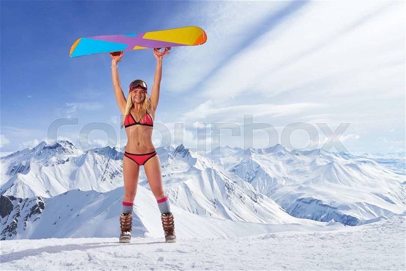 Smiling unrecognizable blonde girl in bikini swimsuit with snowboard in outstretched arms above head against of snowy mountains in sunlight, stock photo