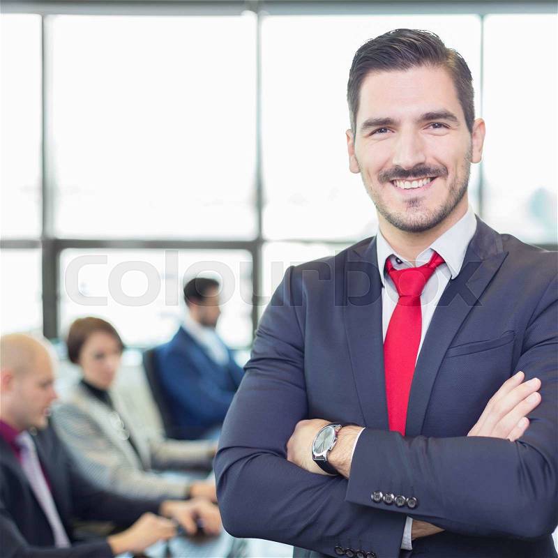 Successful team leader and business owner proudly standing with crossed arms with coworkers working in office in background. Business and entrepreneurship concept, stock photo