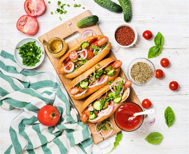 Homemade hot-dogs on wooden serving board with fresh vegetables, spices, ketchup and mustard over white painted old wooden background, top view, stock photo