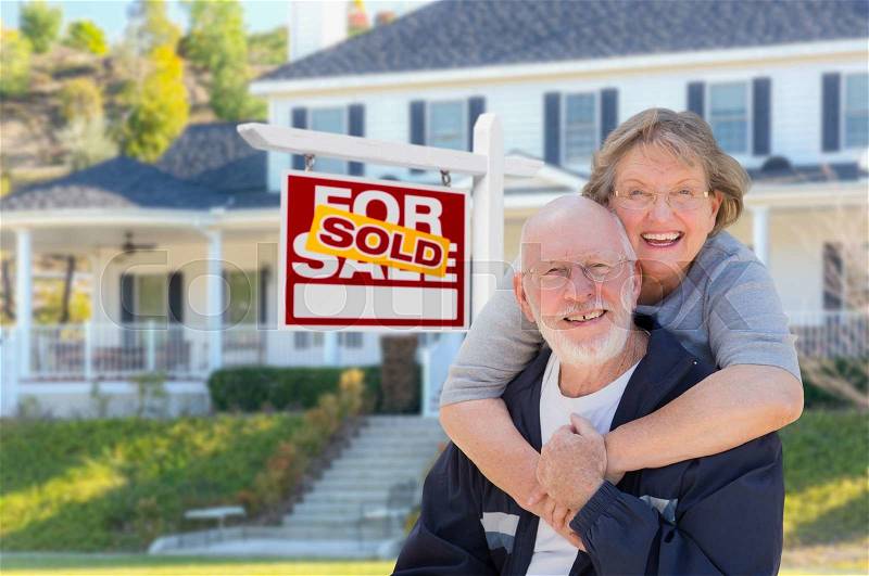 Senior Adult Couple in Front of Sold Home For Sale Real Estate Sign and Beautiful House, stock photo