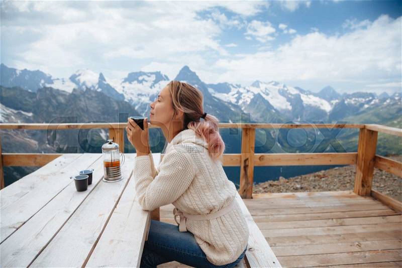Woman drinking warm tea in the rustick wooden outdoor cafe on mountain, alpine view, snow on hills. Dombay, Karachay-Cherkessia, Caucasus, Russia, stock photo