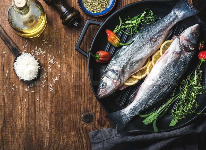 Ingredients for cookig healthy fish dinner. Raw uncooked seabass with rice, olive oil, lemon slices, herbs and spices on black grilling iron pan over rustic wooden background, stock photo