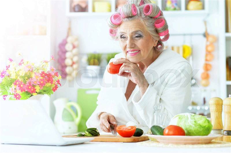 Senior woman in hair rollers at kitchen with laptop and vegetables, stock photo